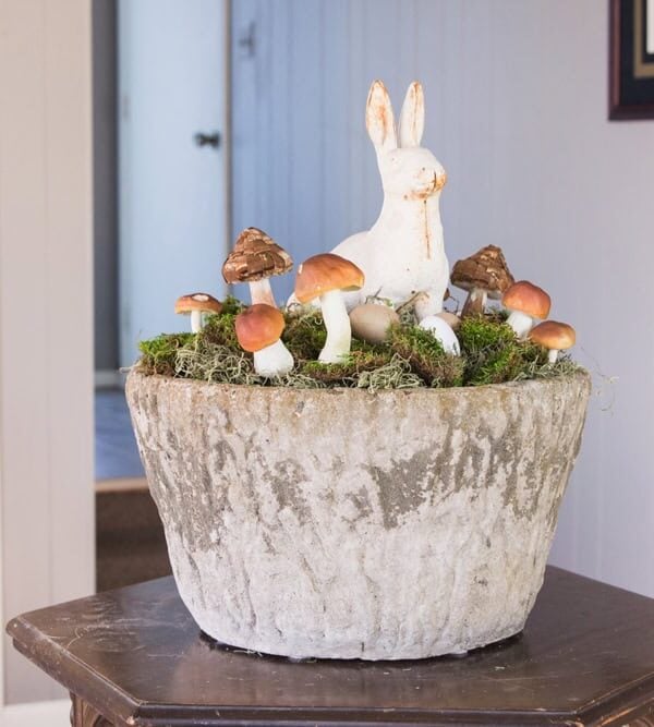 Concrete large vessel sitting on a wooden table decorated with cast iron bunny and small mushrooms for an Easter display.