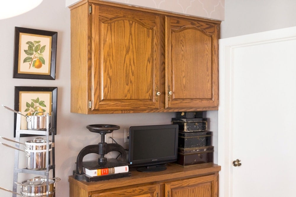 Cherry brown cabinets with television, cash boxes and weight sitting on kitchen countertop along with kitchen pot tree