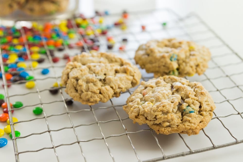 Three monster cookies sitting on cooling rack with lots of M&M's and cookie jar in background