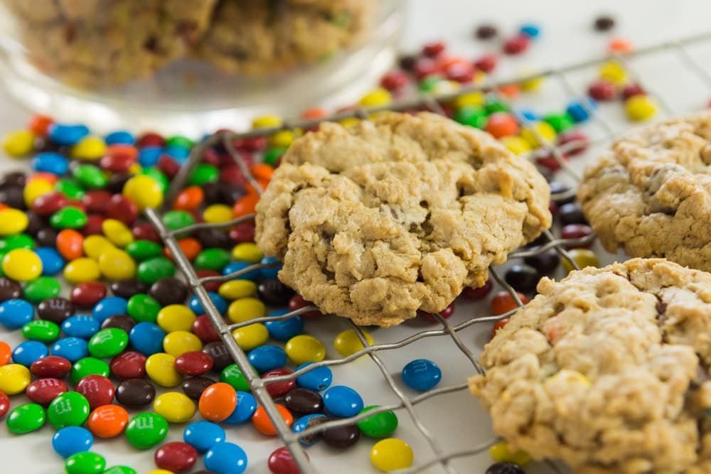 Monster cookie made from oats sitting on cooling rack with two other cookies with M&M's sprinkled underneath rack with glass cookie jar in background