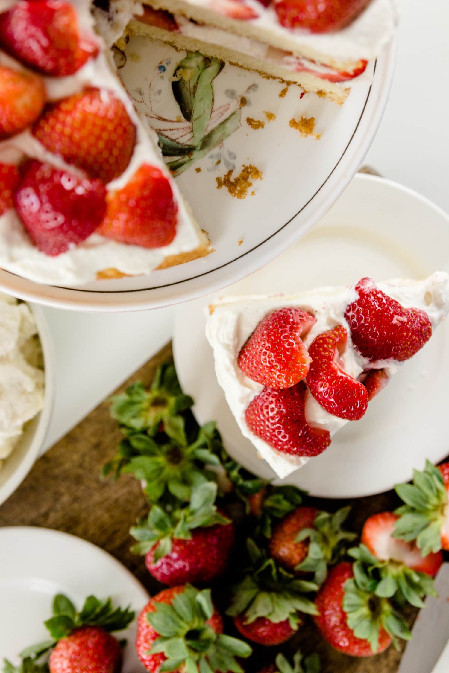 Top down view of strawberries and whipped cream on top of a cake plate with cutting board with strawberry tops and cut piece of cake on a plate