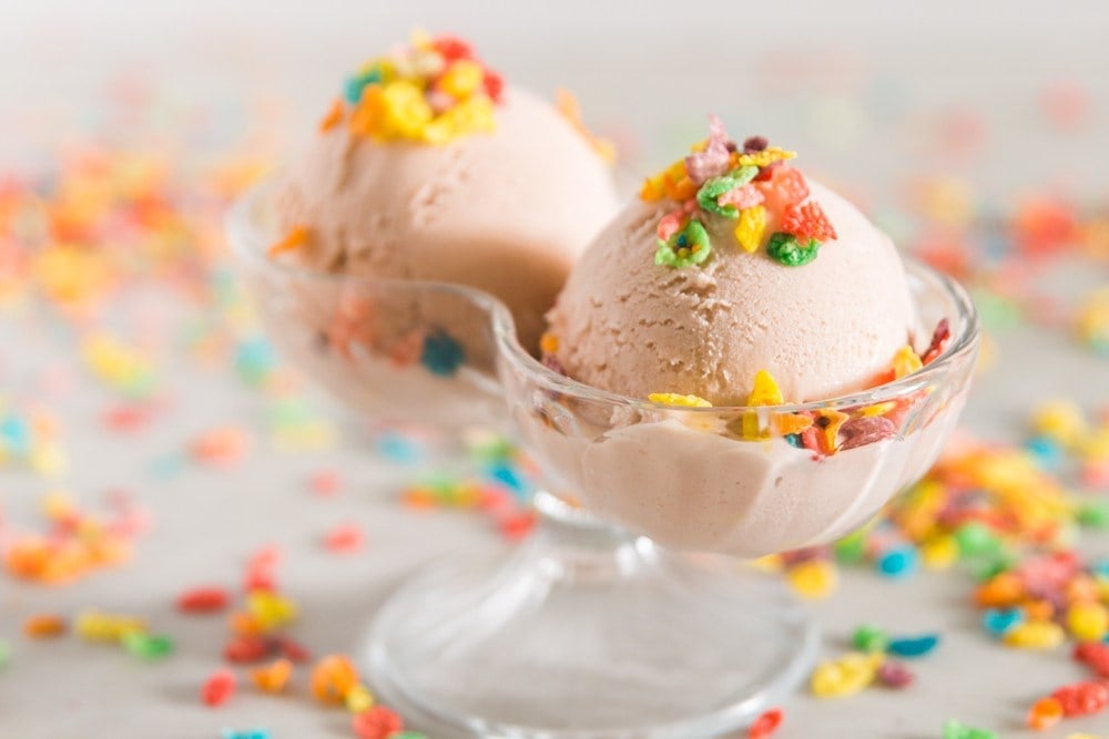 Fruity Pebbles cereal sprinkled on top of pink ice cream in a double-scoop bowl with Fruity Pebbles cereal in the background