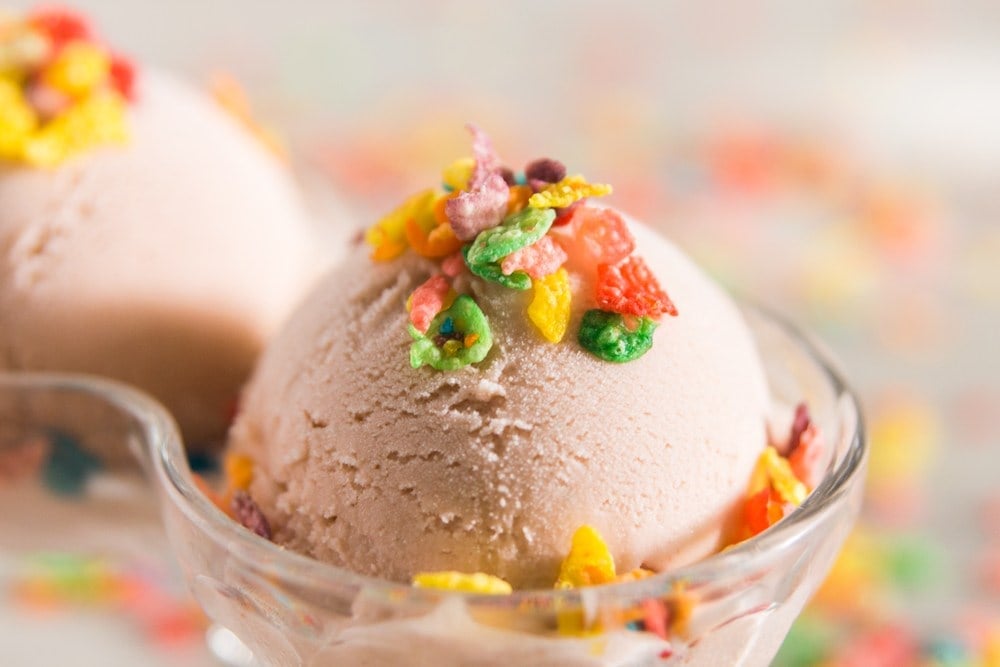 Fruity Pebbles cereal sprinkled on top of pink ice cream in a double-scoop bowl with Fruity Pebbles cereal in the background
