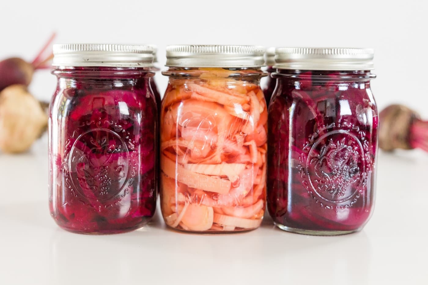 Three different kinds of canned beet recipes in Ball canning jars on a white surface with red and yellow beets in the background.