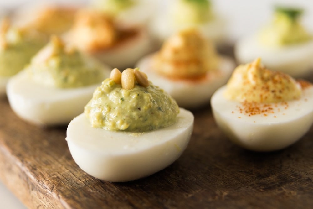 Hardboiled egg sitting on wood board with green basil pesto filling topped with pine nut.