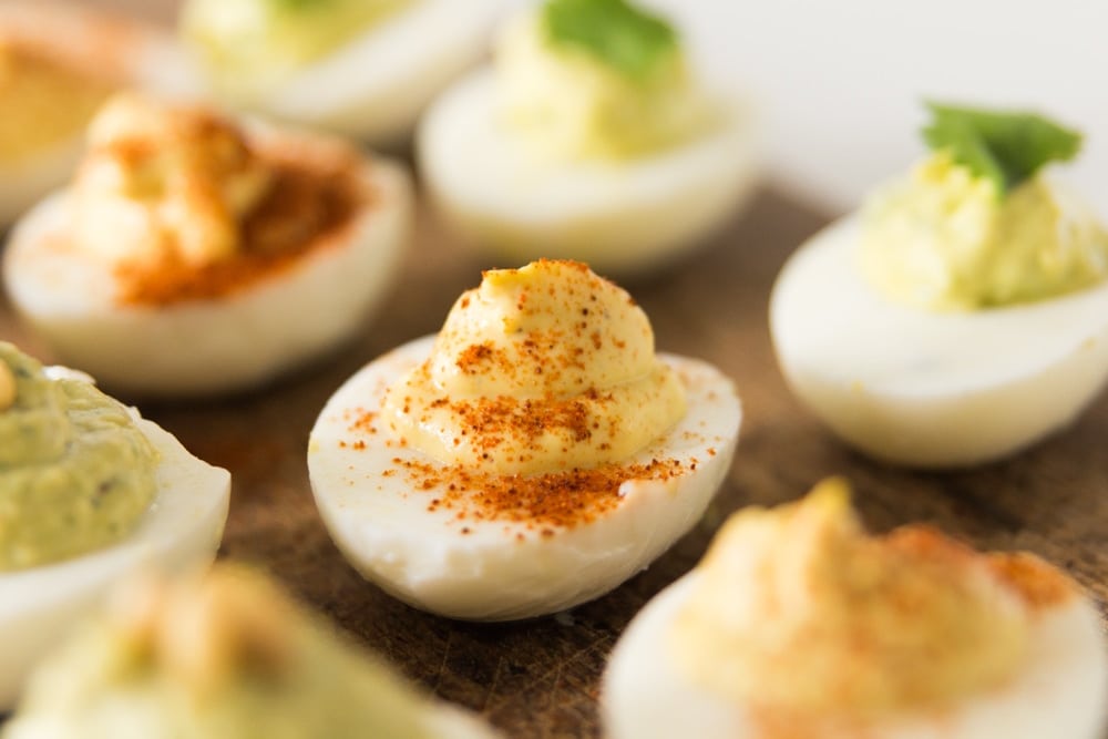 Classic hardboiled egg sitting on wood board with different types of eggs sitting in front of and behind with egg filling topped with paprika.