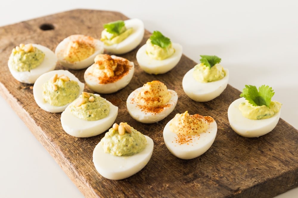 Twelve hardboiled eggs sitting on wood board with four of each kind topped with pine nuts, paprika, and cilantro.