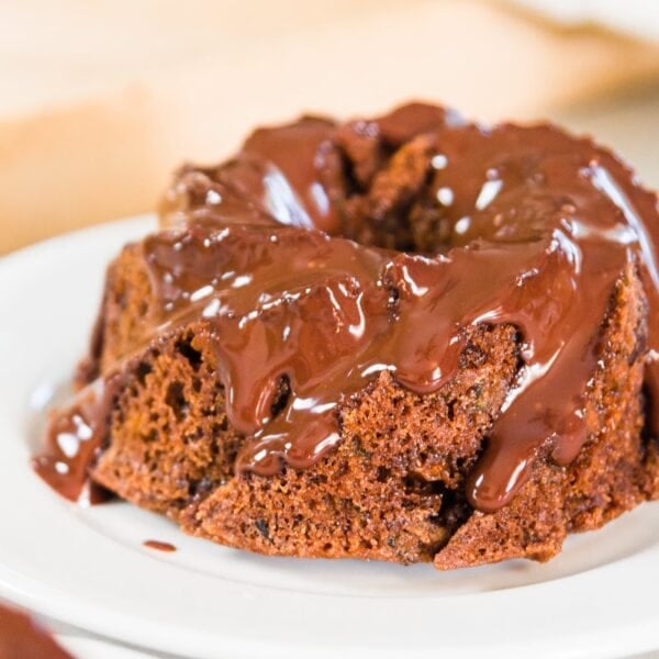 Small Bundt chocolate zucchini cake with chocolate ganache poured over top sitting on white plate with cutting board in background