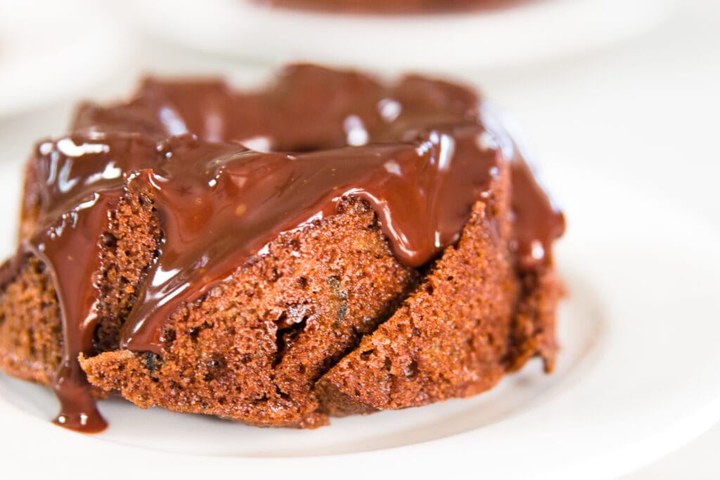 Close up view of brown colored chocolate zucchini cake dripping with chocolate frosting made from ganache sitting on white plate