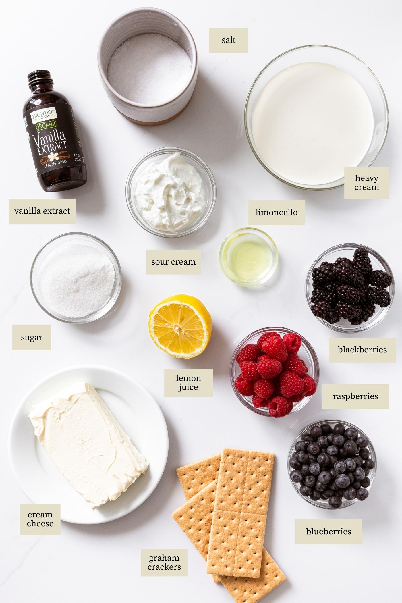 White countertop filled with lots of ingredients to make triple berry cheesecake include graham crackers, berries, cream, lemon, and vanilla extract.