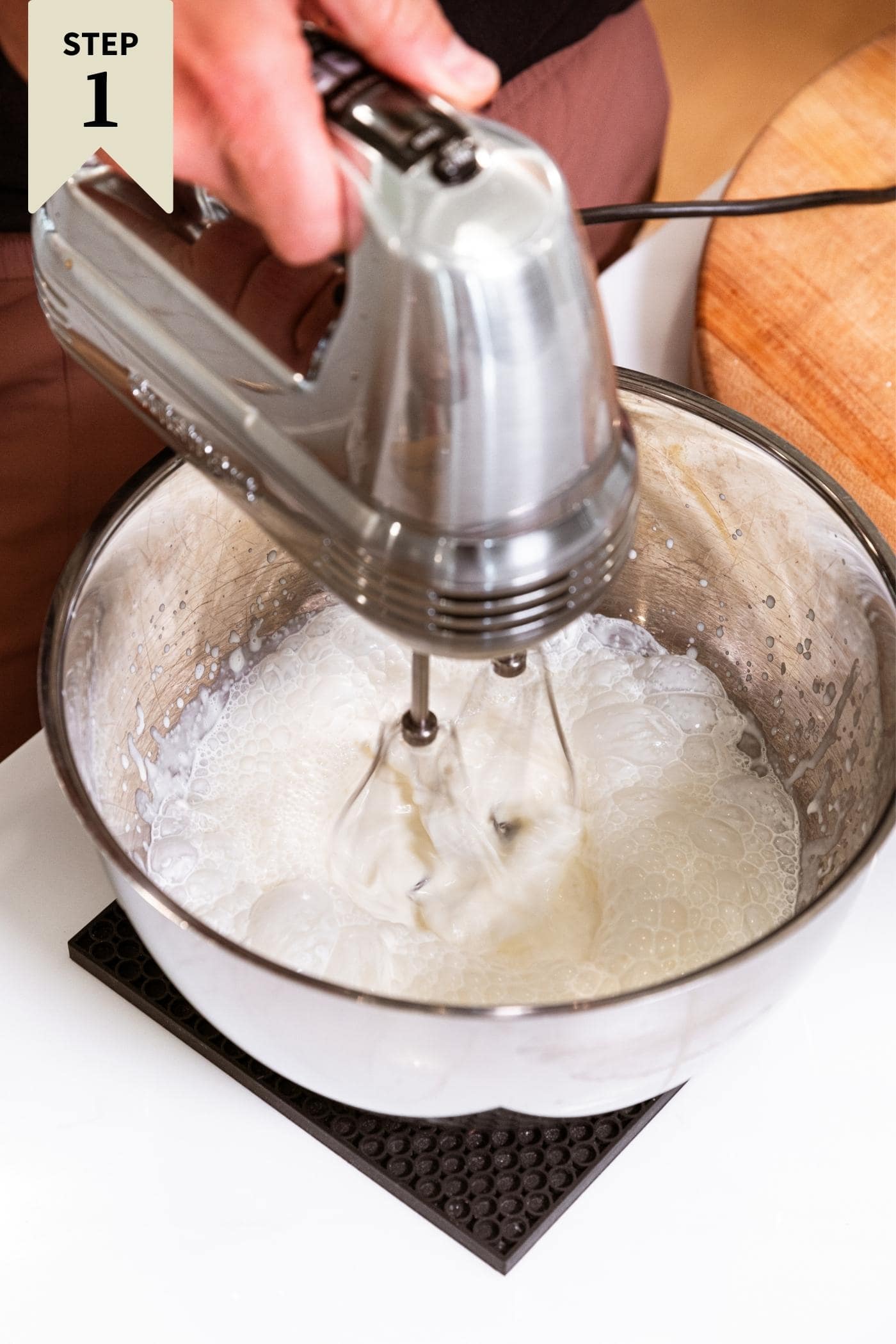 Hand holding handheld mixer mixing up whipped cream in steel bowl sitting on white countertop.