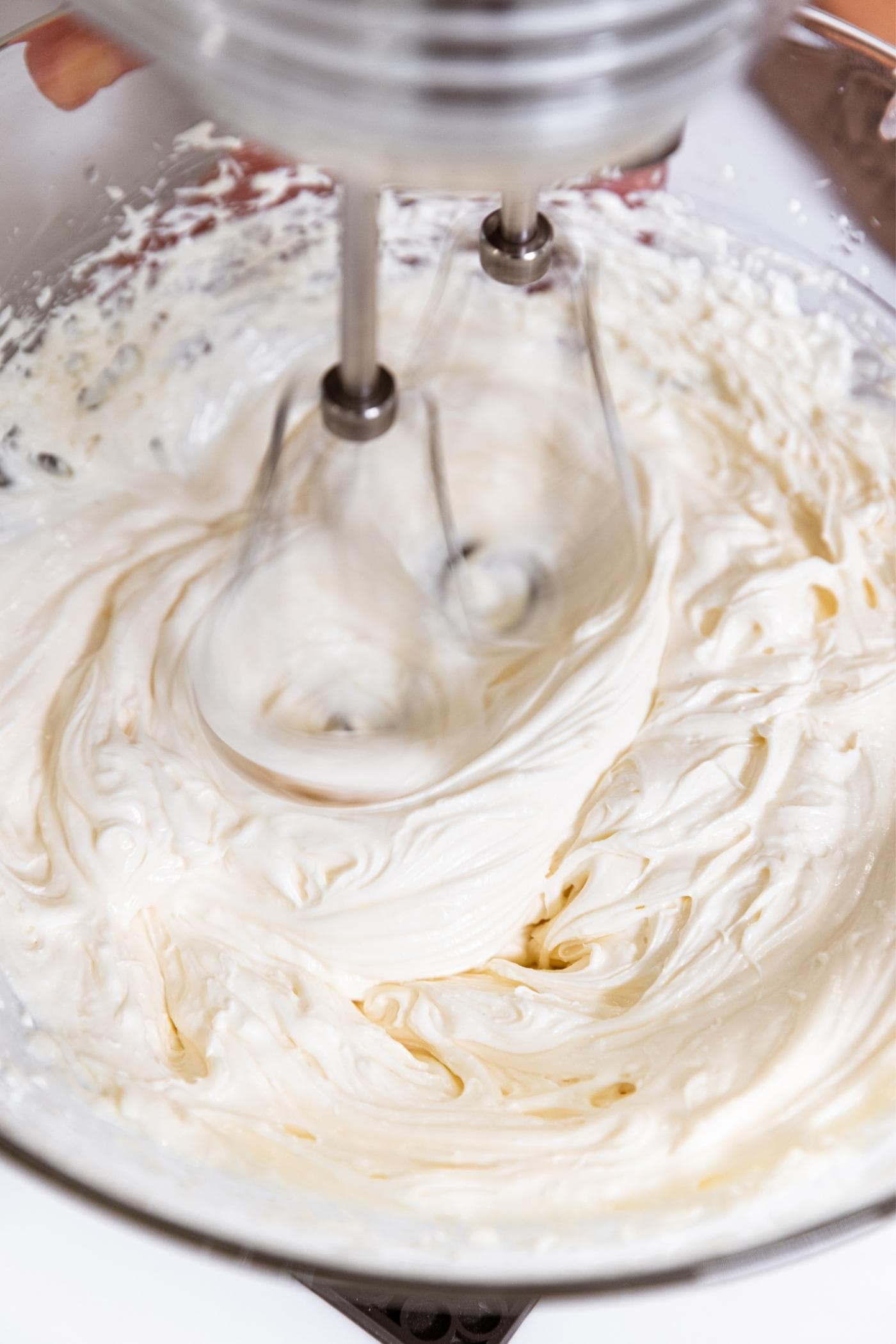 Handheld mixer spinning cream cheese in glass bowl on white countertop.