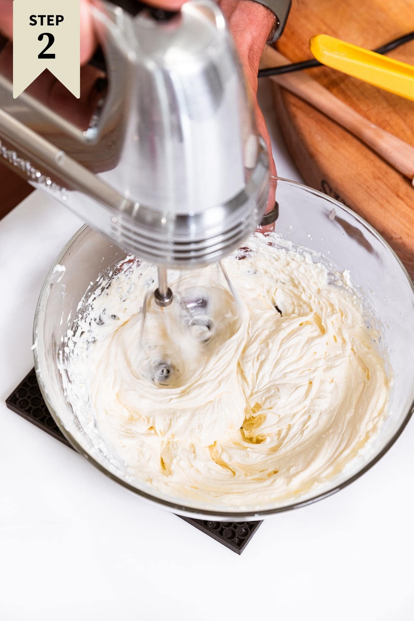 Hand holding handheld mixer mixing up cream cheese in glass bowl sitting on white countertop.