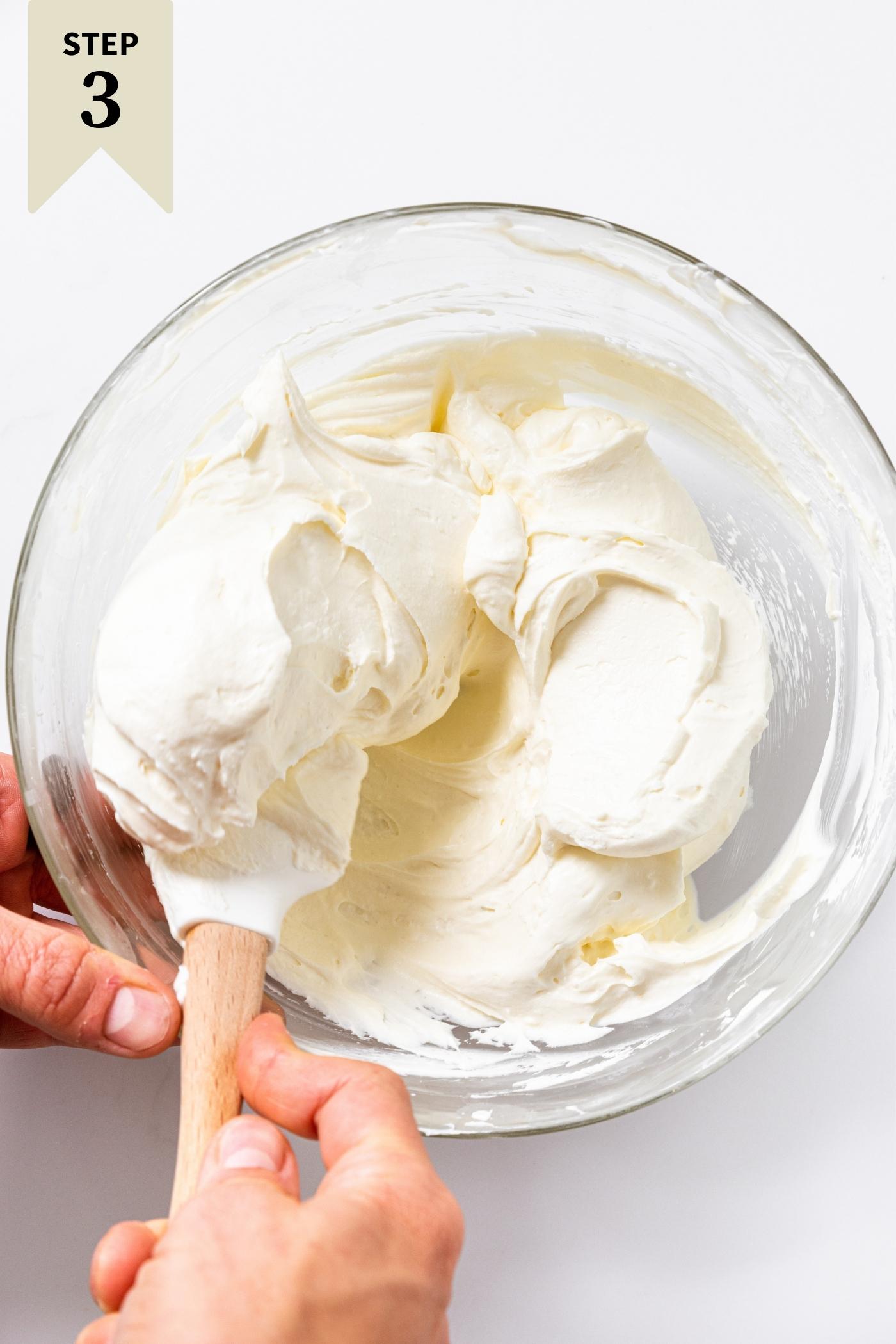 Glass bowl filled with cream cheese and whipped cream mixture being folded by hand holding white spatula all on white countertop.