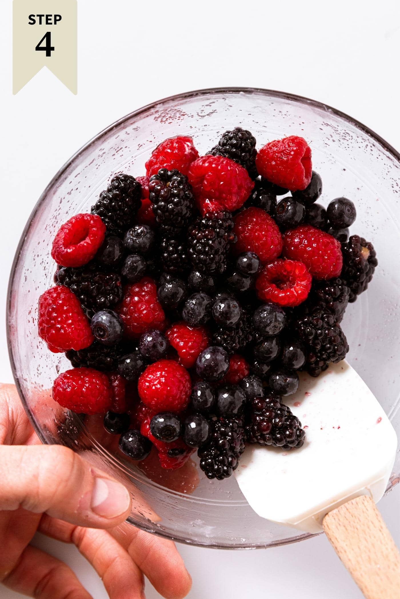 Glass bowl sitting on white countertop filled with three types of berries being mixed by white spatula being held.