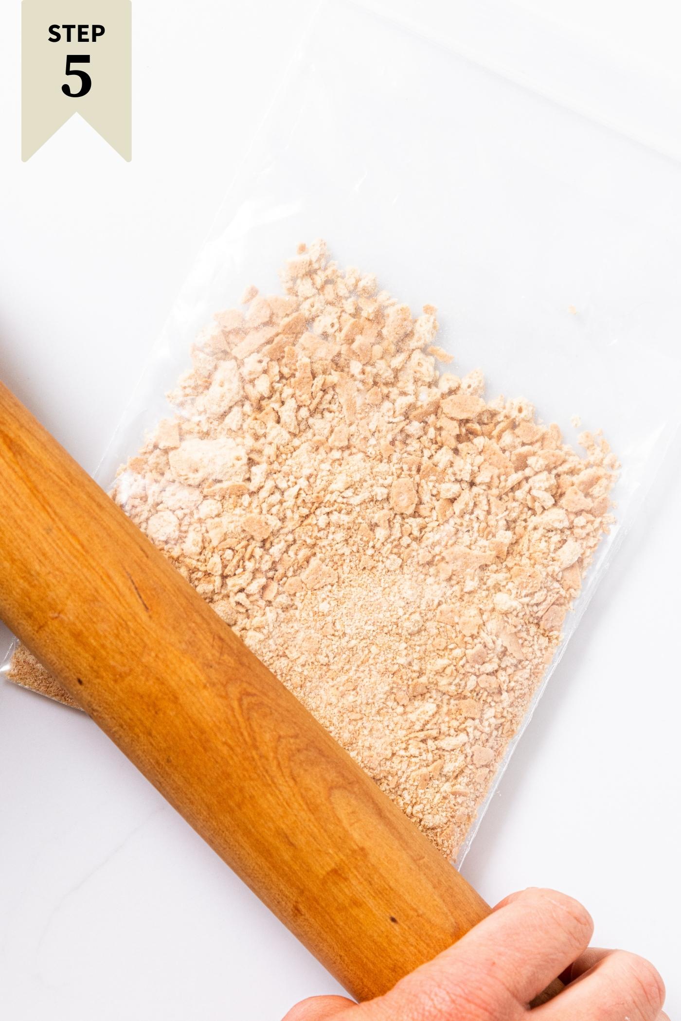 Wooden rolling pin sitting on plastic bag with crushed up graham crackers sitting on white countertop.