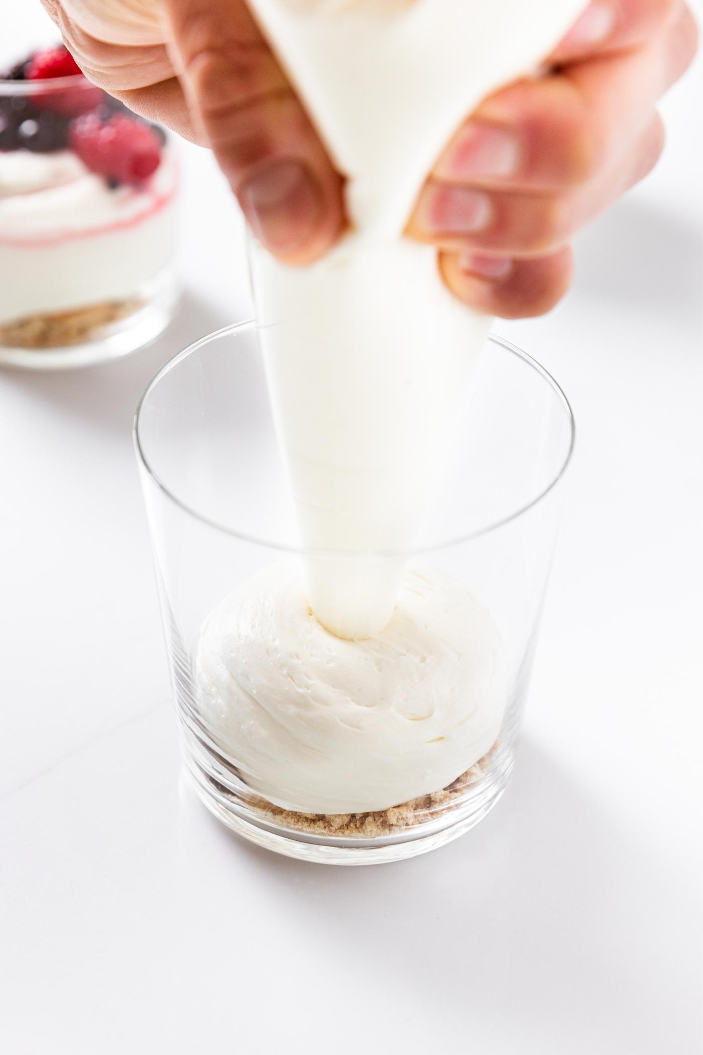Hand piping cream cheese mixture into glass with graham cracker crumbs on bottom sitting on white countertop.