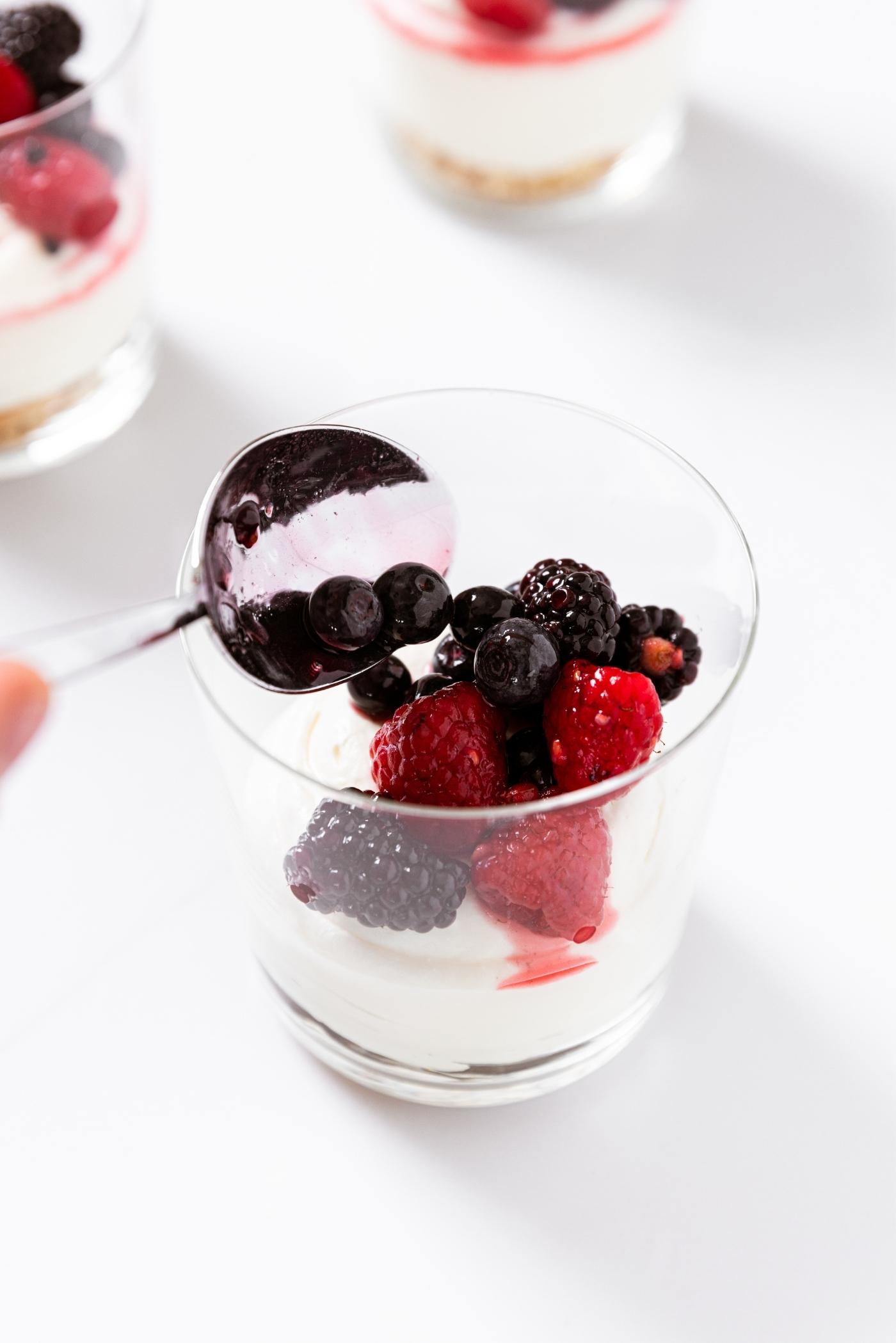Hand spooning mixed berry mixture on top of cream cheese mixture in glass sitting on white countertop.