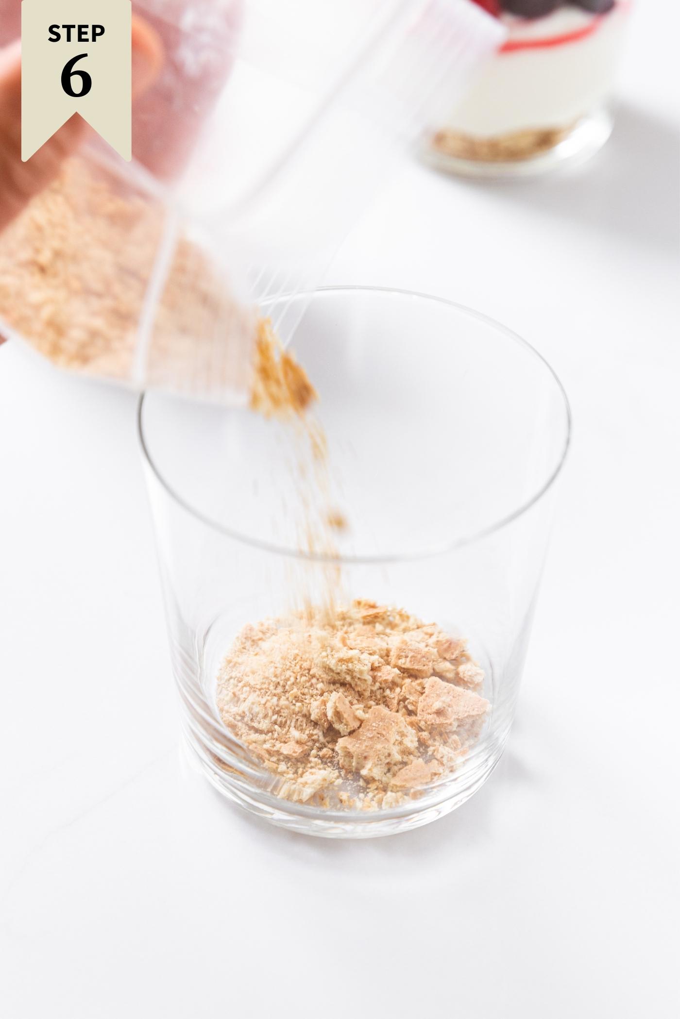 Hand pouring graham cracker crumbs into glass sitting on white countertop.
