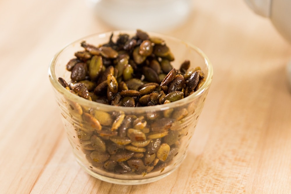 Small glass bowl with roasted pepitas sitting on light wood surface with white bowl in background