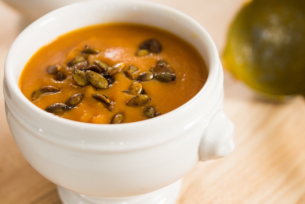 White bowl filled with orange colored chipotle squash soup with pepitas sprinkled over the top with ladle in background on wood surface
