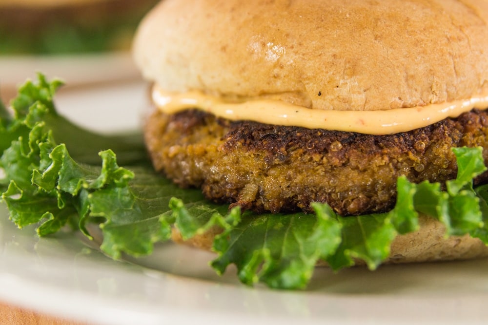 Close up view of side of quinoa squash burger sitting on bed of lettuce with chipotle mayonnaise slathered on top with bun capping the entire thing