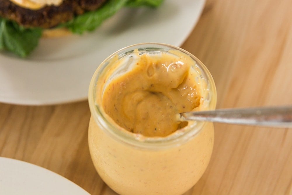 Glass container filled with orange colored chipotle mayonnaise with spoon sticking out all on wood surface with burger in background