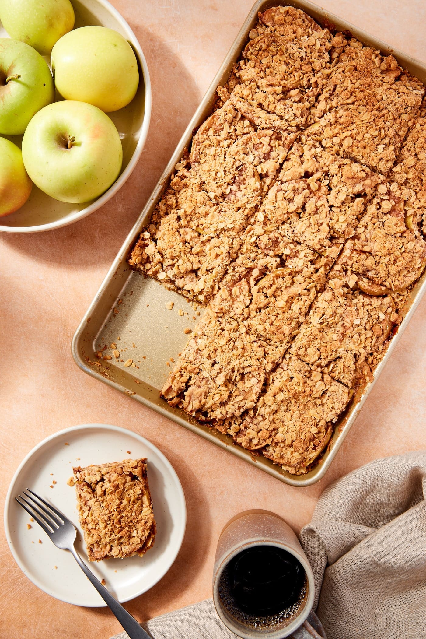 Gold baking pan filled with baked apple pie bars sliced into rectangles with slice sitting on a white plate with bowl of apples.