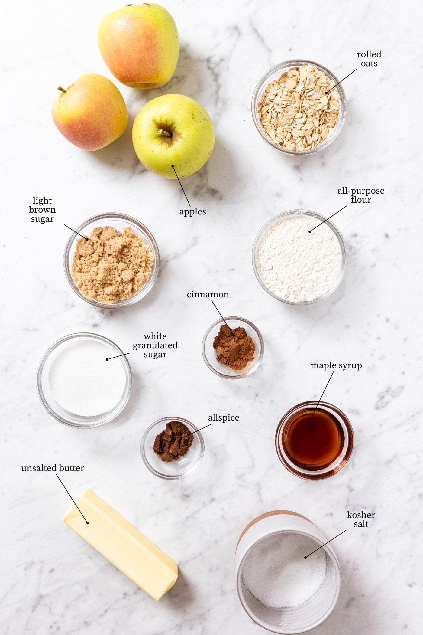 White marble surface filled with all ingredients needed to make apple pie bars including sugars, apples, oats, maple syrup, spices, and more.