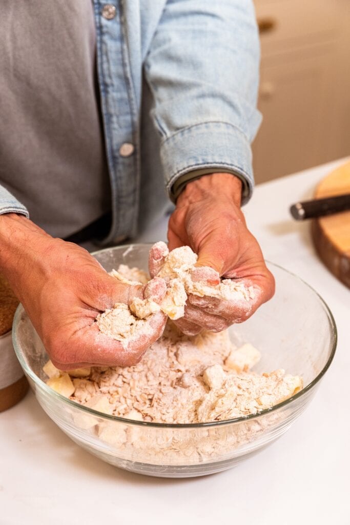 Two hands pressing butter with flour mixture in large glass bowl on white surface.