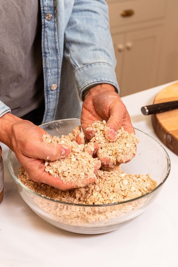 Two hands crumbling together butter with oats and flour in a glass bowl for apple pie bars.