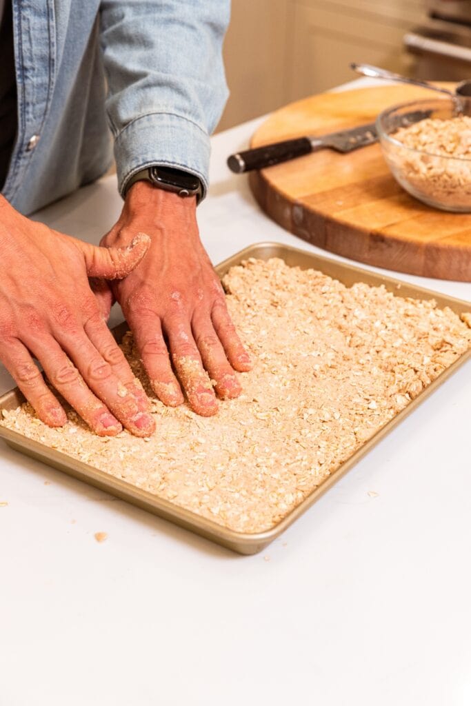 Two hands pressing crust for apple pie bars into a large gold baking pan sitting on white surface.
