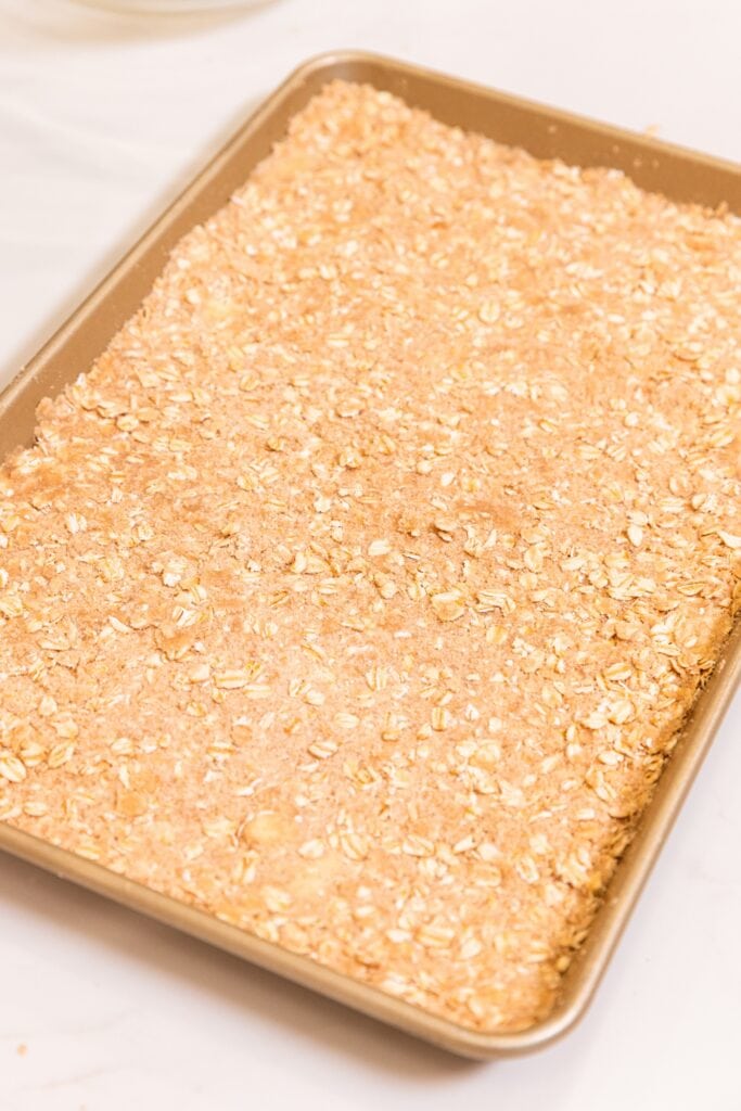 Gold baking pan filled with crust made with oats and brown sugar sitting on white surface before going into the oven.