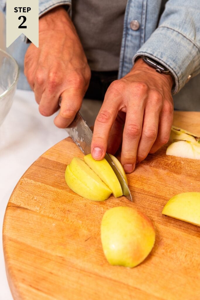 Two hands using a knife to slice up yellow apples on a wooden cutting board.