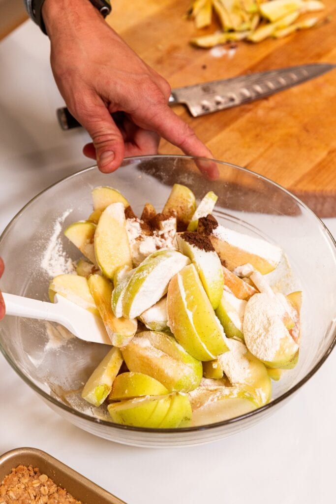 Glass bowl filled with sliced apples with sugar and spices on top all on white surface.
