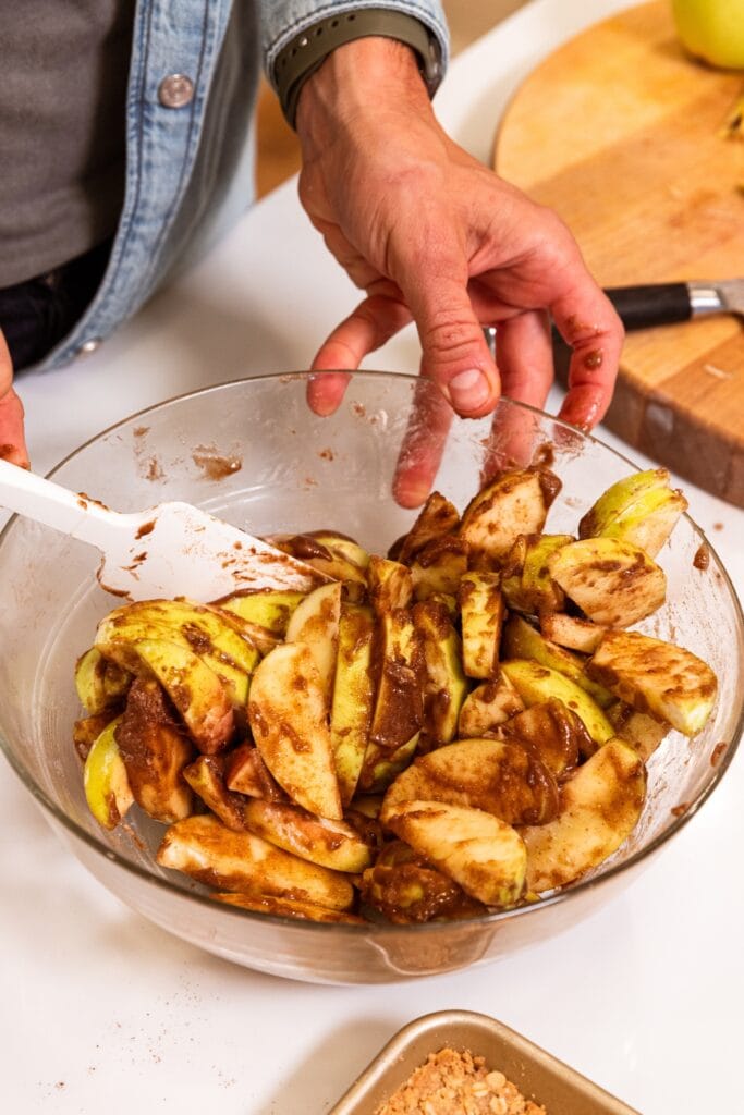 Glass bowl filled with apple slices covered in cinnamon and sugar mixture all on white surface.
