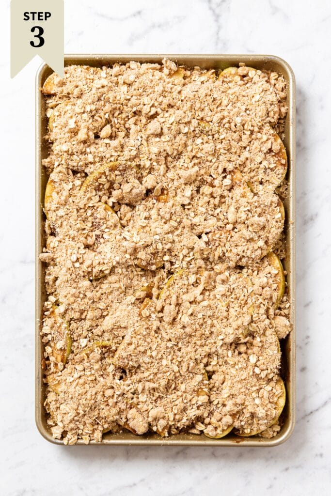 White marble surface with gold baking sheet with apple pie bars covered in streusel topping before being baked.
