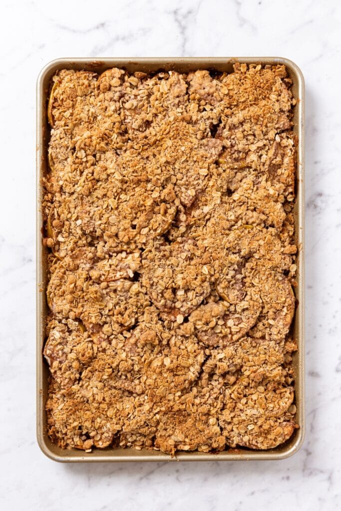 Apple pie bars in gold baking pan right after being baked in the oven all on white marble surface.