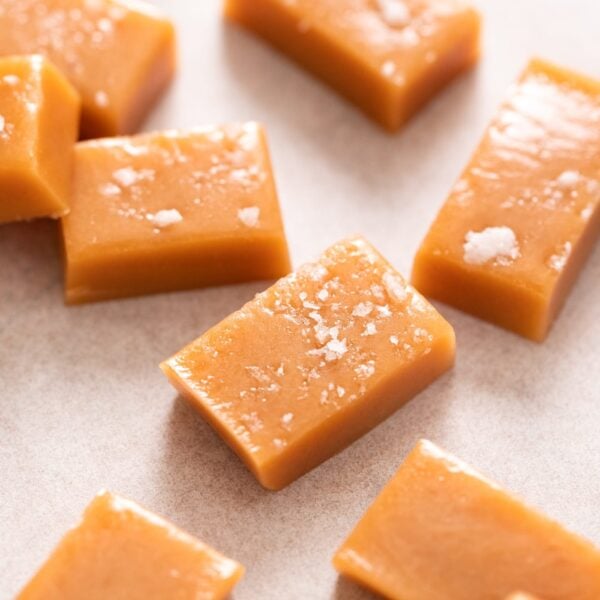 Pieces of golden caramel sitting on white piece of parchment, some topped with flaky sea salt
