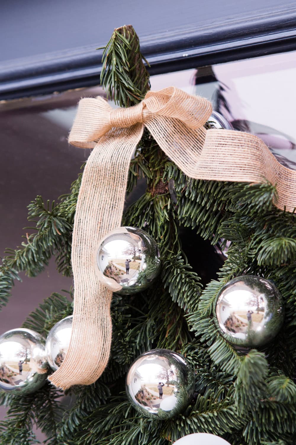 Light taupe colored ribbon tied to the top of an evergreen bundle adorned with silver balls attached to a door.