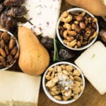 Top down view of three spiced nuts sitting on wood board with chunks of cheese, pears, and dates all around