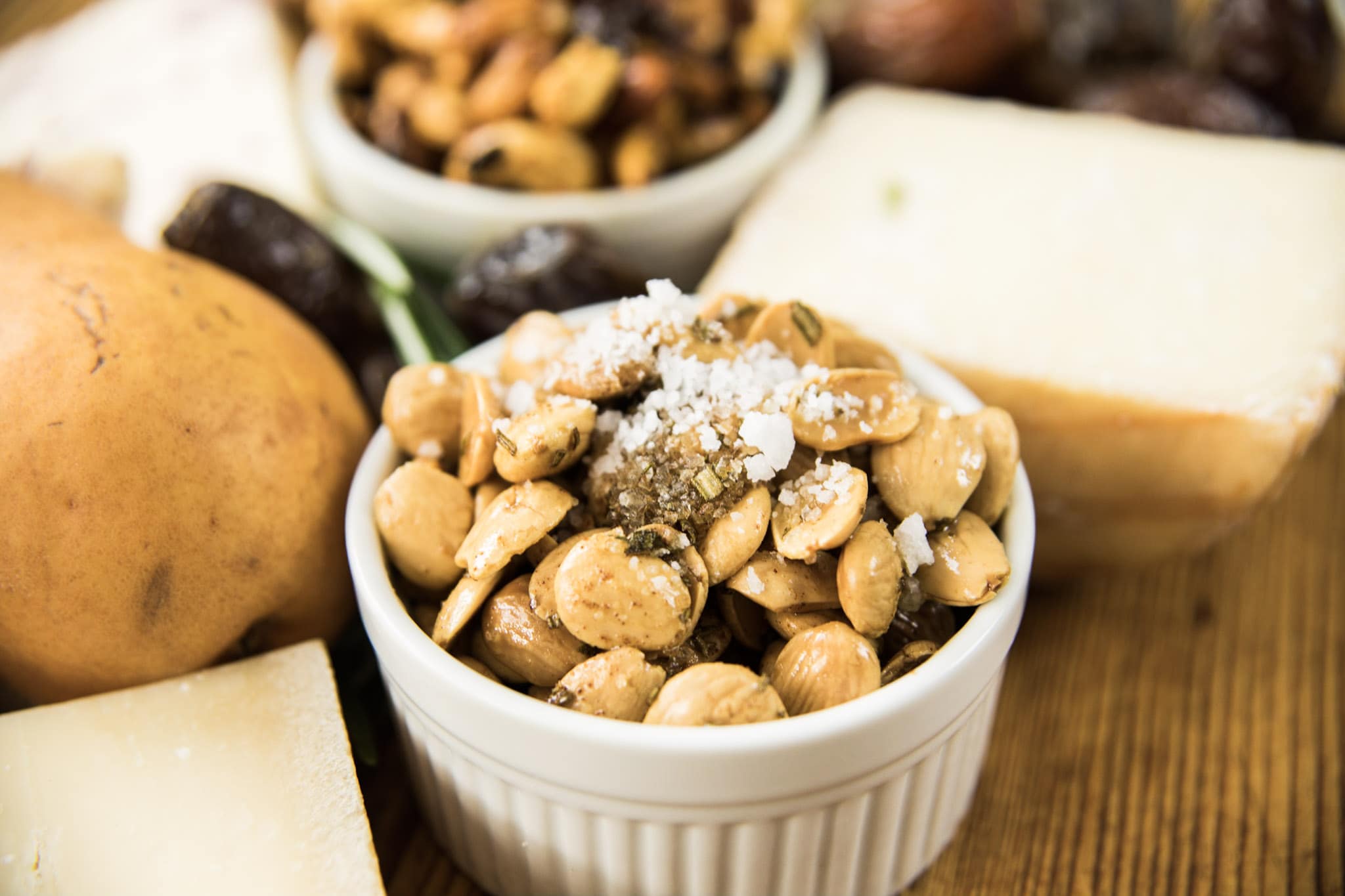Marcona almonds sitting in circular white bowl topped with sea salt with hunks of cheese and pears in background all on wooden board
