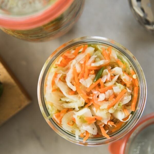 Top down view of glass container filled with curtido relish made from slices of cabbage and carrots with a jalapeño sitting on cutting board beside container