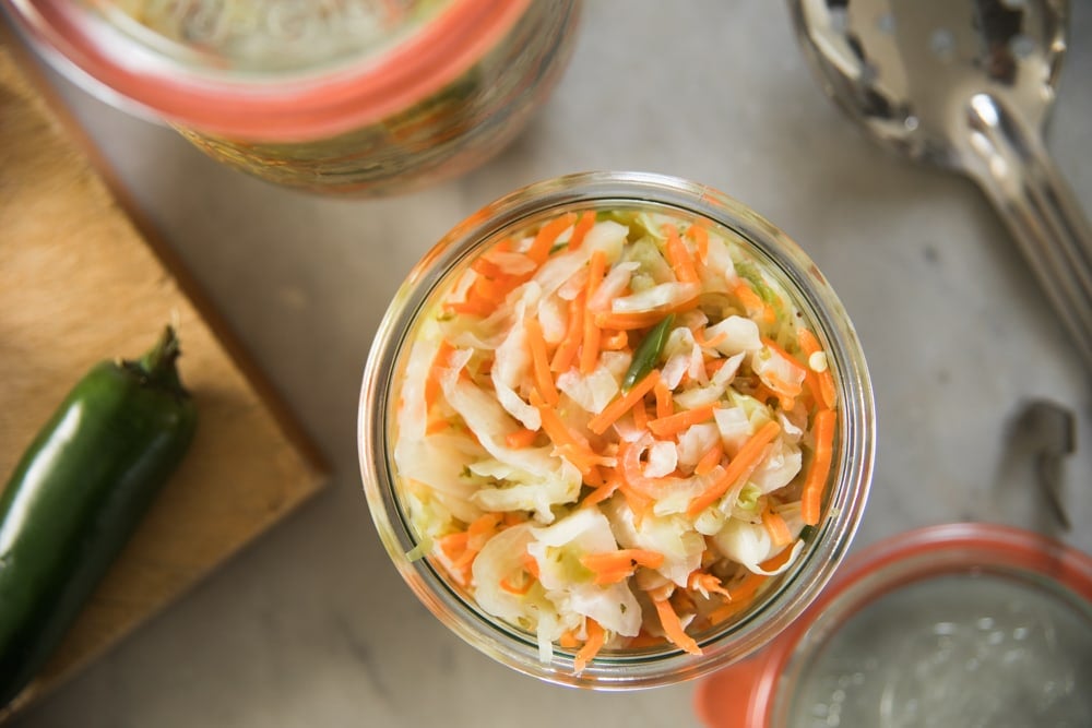 Top down view of glass container filled with curtido relish made from slices of cabbage and carrots with a jalapeño sitting on cutting board beside container