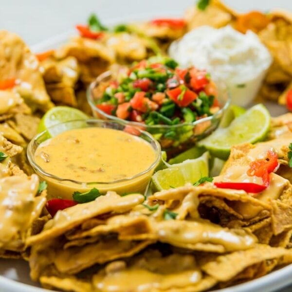 Large white platter filled with tortilla chips and topped with cheese along with containers filled with more cheese, pico de gallo, and sour cream all on marble surface
