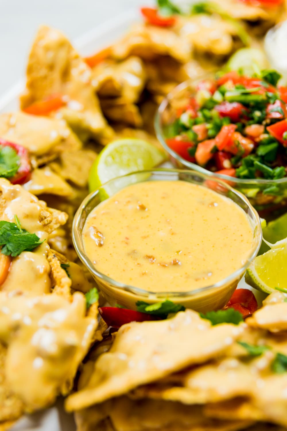 Ring of stadium nachos topped with cheese mixture with glass container filled with cheese dip and another container of pico de gallo