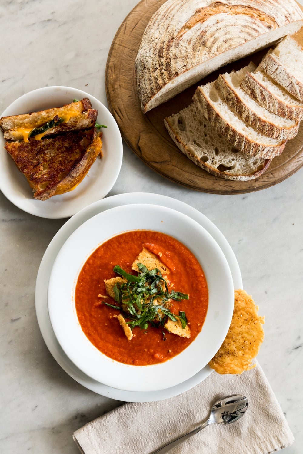 Top down view of marble surface with bowl of tomato and roasted red pepper topped with Parmesan crisps and strips of basil on top of soup with grilled cheese on plate beside