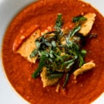 Top down view of white bowl filled with red colored tomato and roasted red pepper soup with Parmesan crisp and strips on basil on top