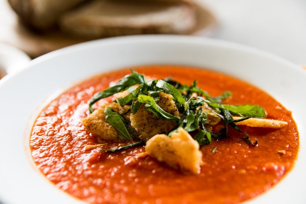 White bowl filled with red colored tomato and roasted red pepper soup with Parmesan crisps and strips of basil on top