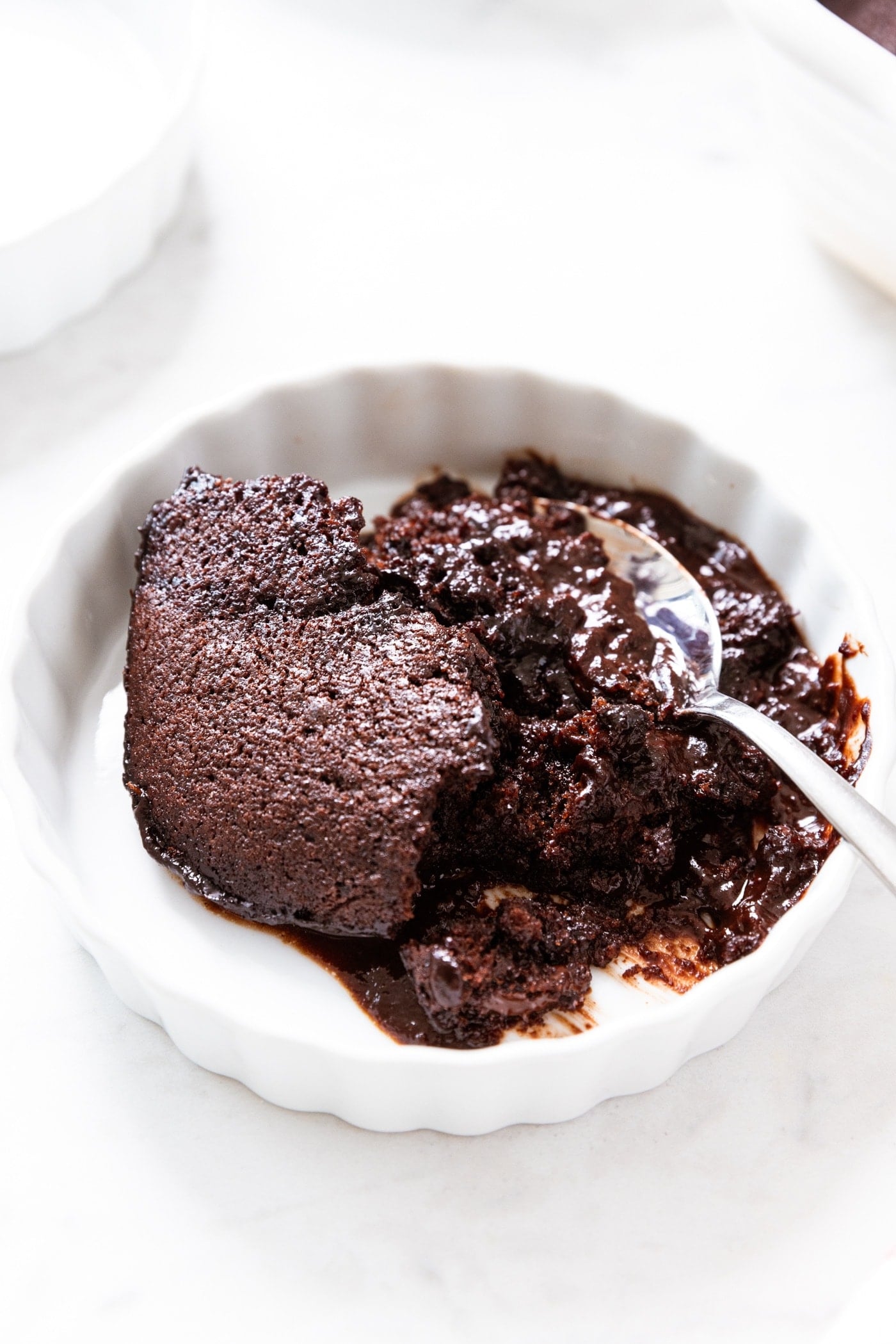 Round white bowl filled with chocolate fudge pudding cake with spoon resting against the side before scooping up a bite.